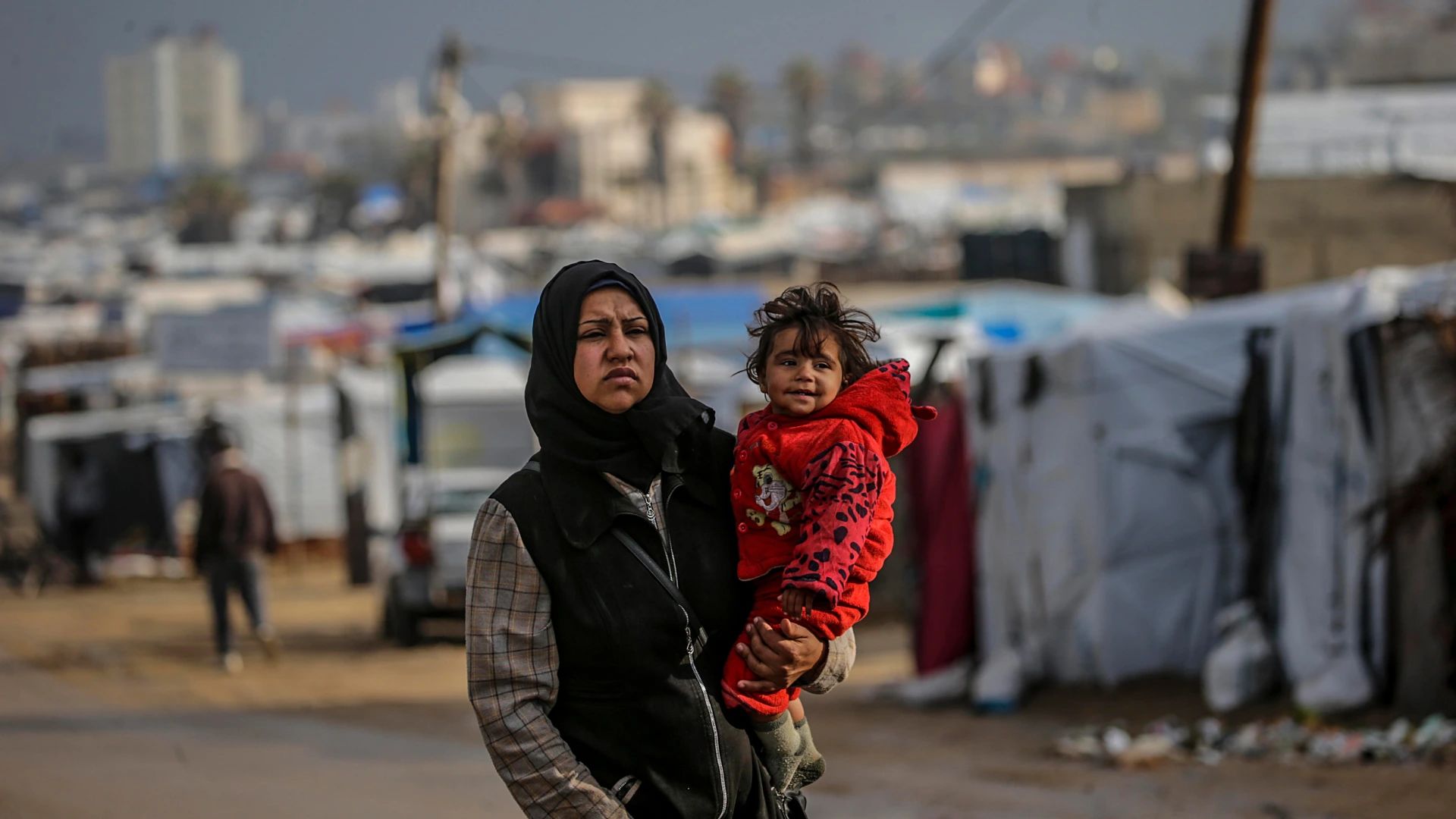 Deir Al Balah (Gaza), 30/12/2024.- Una dona palestina desplaçada interna, portant la seva filla, camina entre tendes en un dia ventós a l’oest de Deir Al Balah, al centre de la Franja de Gaza. Segons l’ONU, almenys 1,9 milions de persones (nou de cada deu) a Gaza són desplaçades internes, incloent-hi persones desplaçades repetidament. Des de l’octubre del 2023, només un 11% de la Franja de Gaza no ha estat afectada per ordres d’evacuació emeses per Israel, segons l’OCHA. EFE/EPA/MOHAMMED SABER